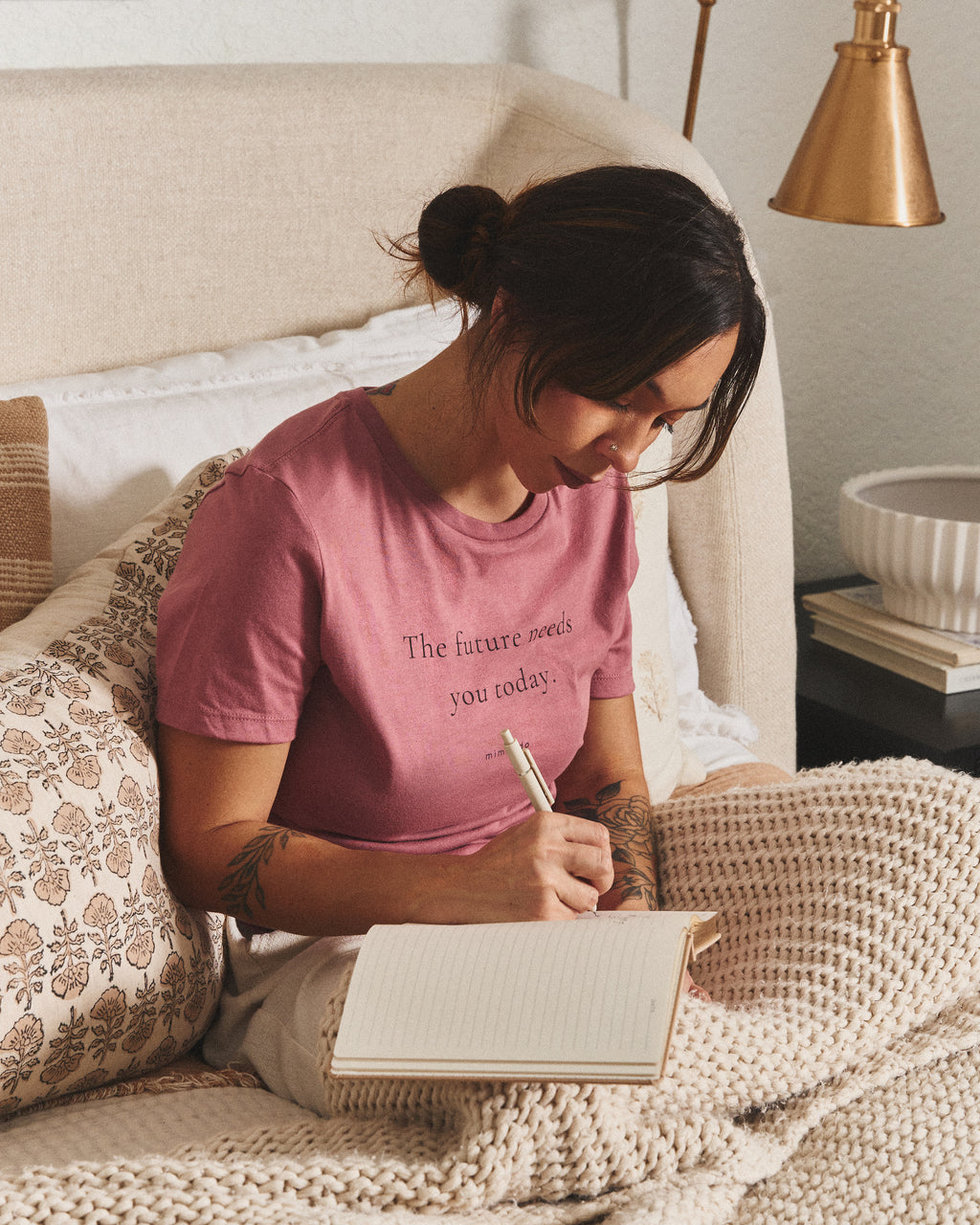A woman writes in a notebook on a bed, in the Future Tee. The print reads: The future needs you today.