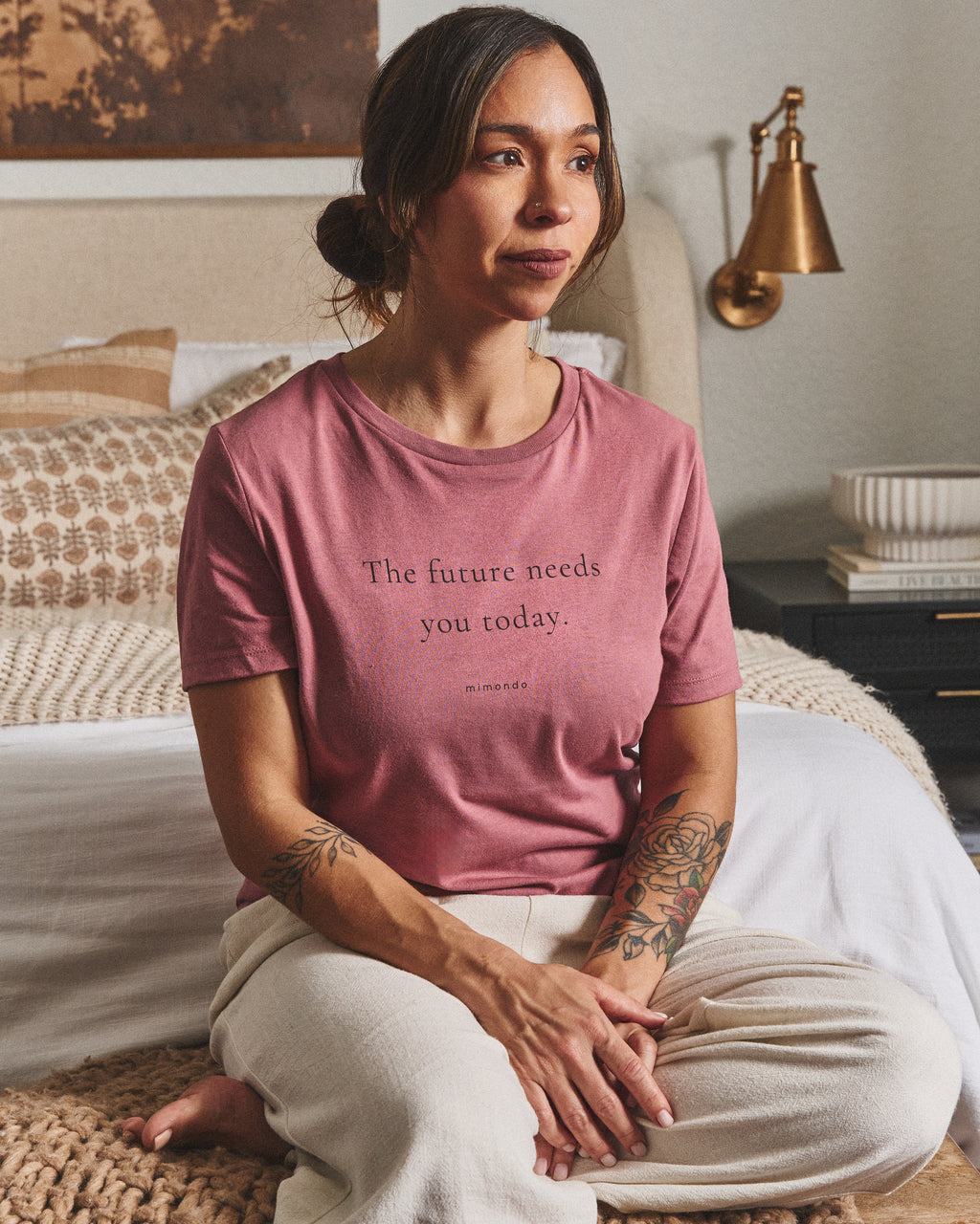 A woman sits on the edge of a bed with her hands in her lap, in the Future Tee. The print reads: The future needs you today.