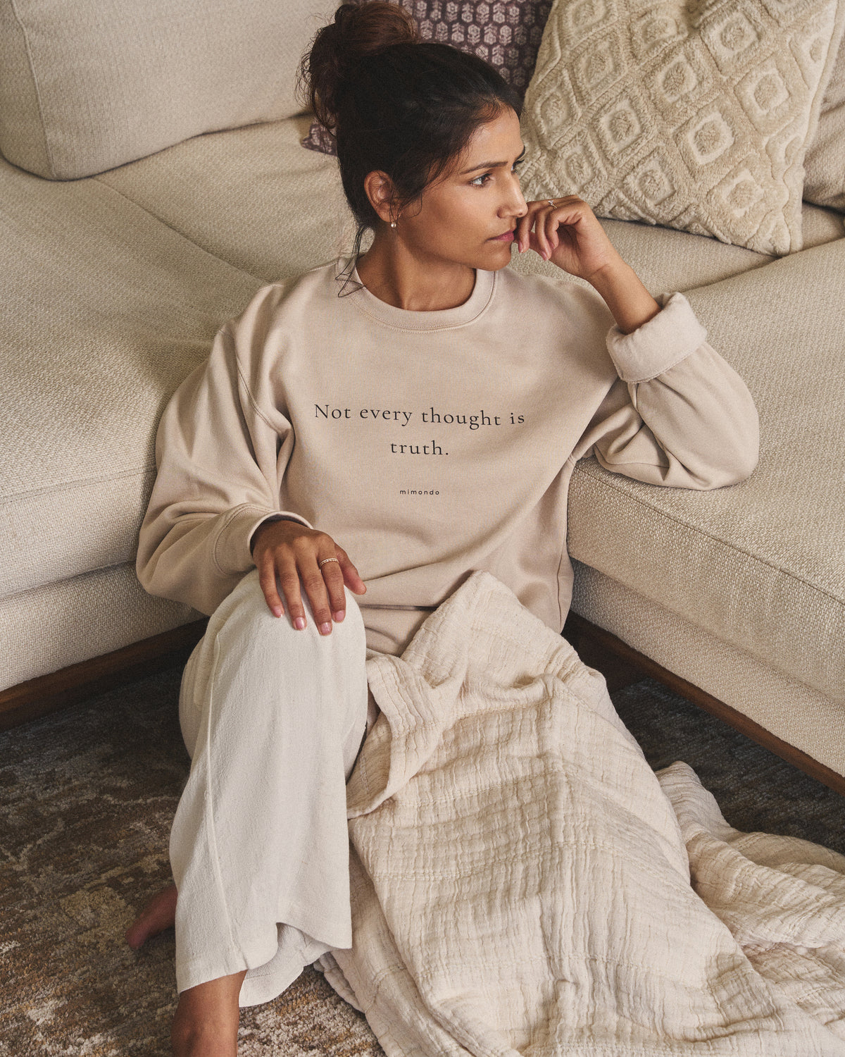 A woman sits on the floor against a sofa, in the Grounded Sweatshirt. The print reads: Not every thought is truth.