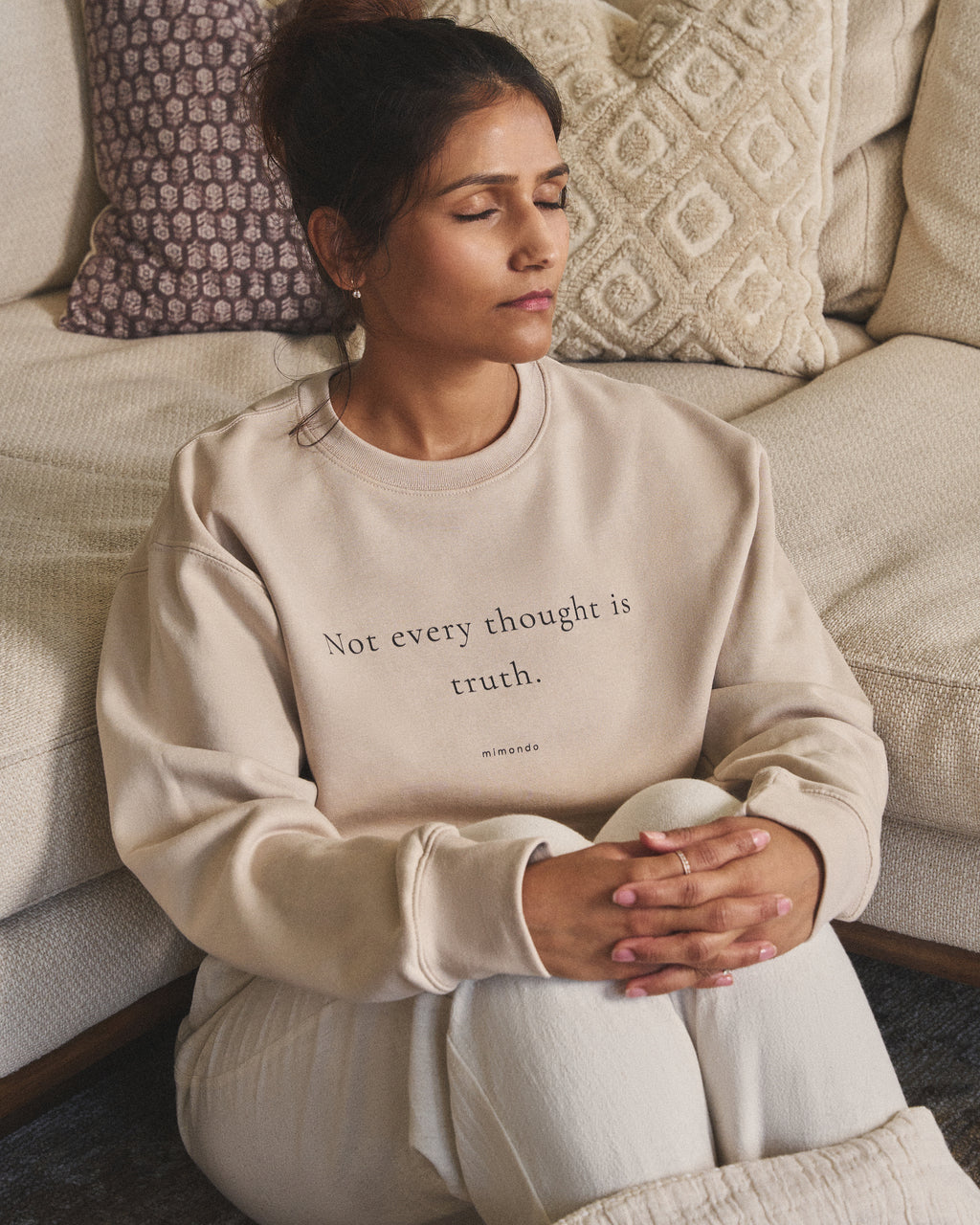 A woman sits with her eyes closed and hands clasped, in the Grounded Sweatshirt. The print reads: Not every thought is truth.