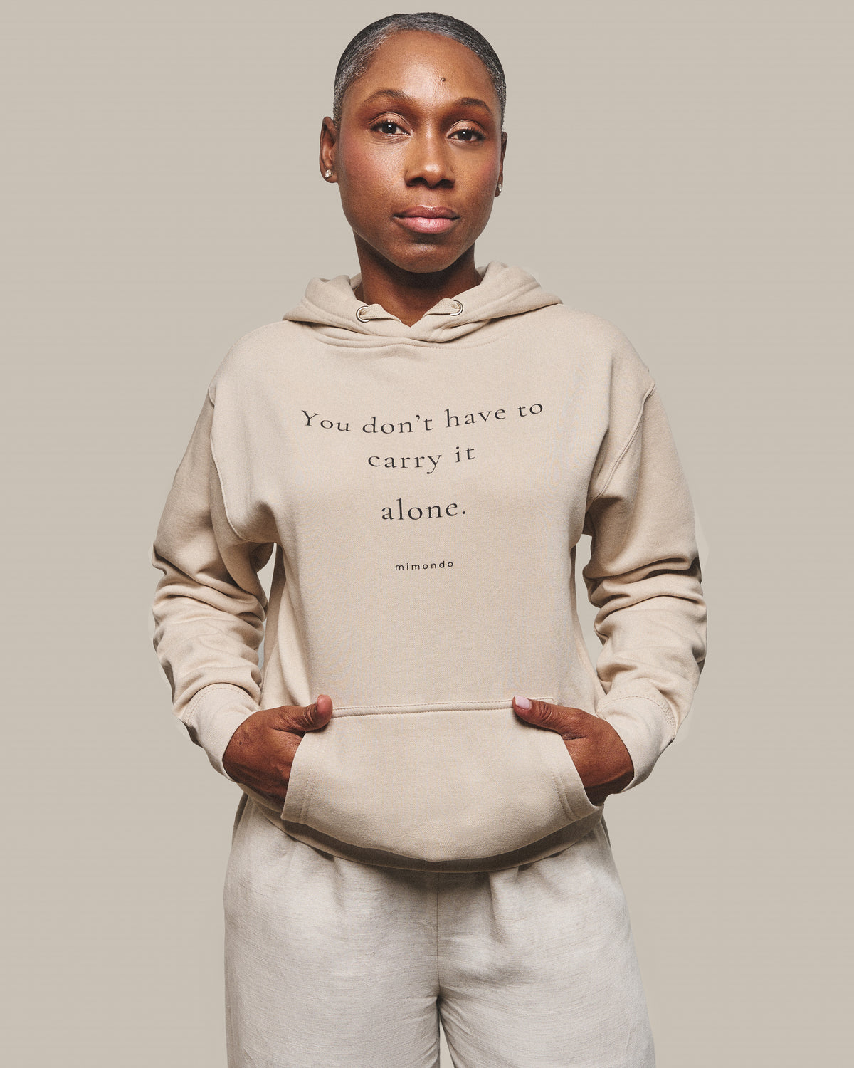 Woman wearing The Hope Hoodie in Bone, hands in the front pocket. The print reads: You don't have to carry it alone.