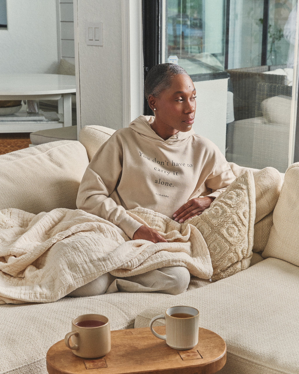 A woman sits on a sofa under a quilt, two mugs nearby, in the Hope Hoodie. The print reads: You don't have to carry it alone.