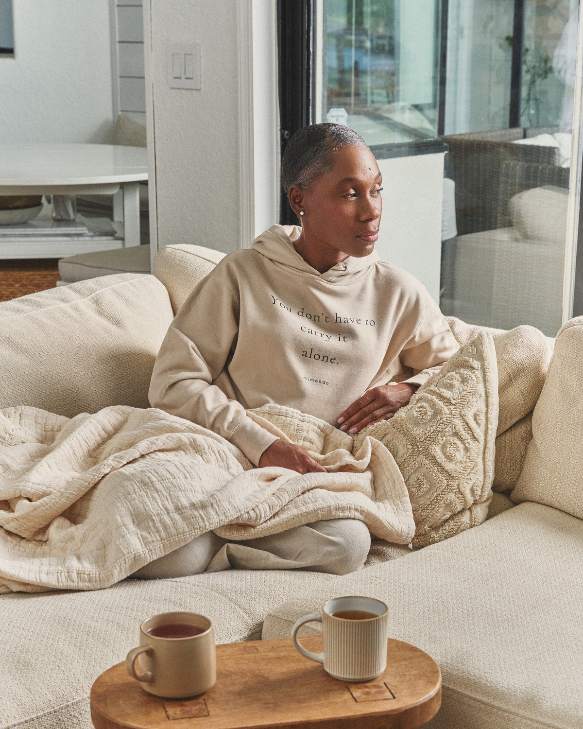 A woman sits on a sofa under a quilt, two mugs nearby, in the Hope Hoodie. The print reads: You don't have to carry it alone.