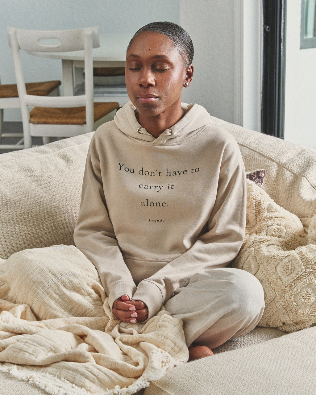 A woman sits cross-legged with her eyes closed, in the Hope Hoodie. The print reads: You don't have to carry it alone.