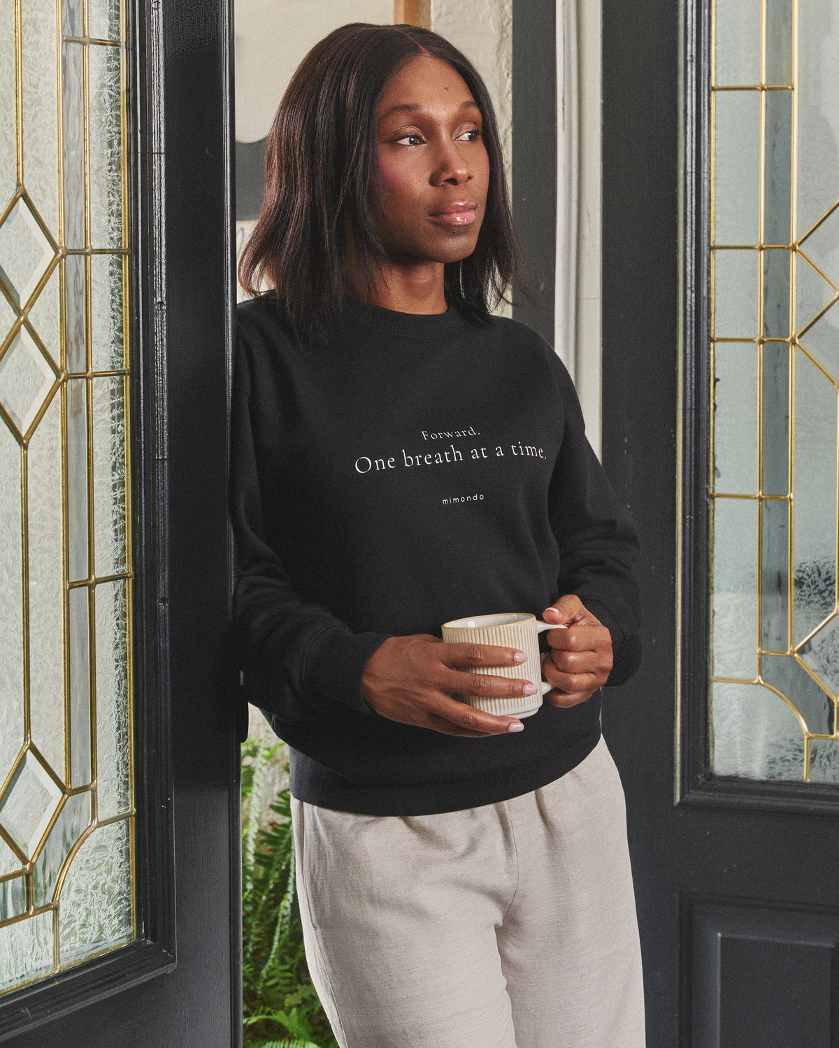 A woman stands in a doorway holding a mug, in the Persist Sweatshirt. The print reads: Forward. One breath at a time.