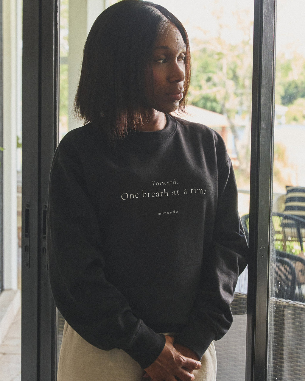 A woman stands at a glass door looking out, in the Persist Sweatshirt. The print reads: Forward. One breath at a time.
