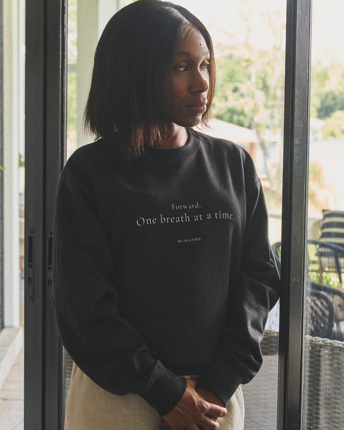 A woman stands at a glass door looking out, in the Persist Sweatshirt. The print reads: Forward. One breath at a time.