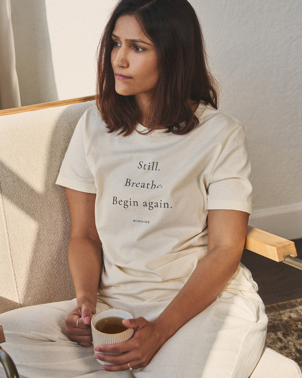 A woman sits cross-legged in a chair holding a cup of tea, in the Still Tee. The print reads: Still. Breathe. Begin again.