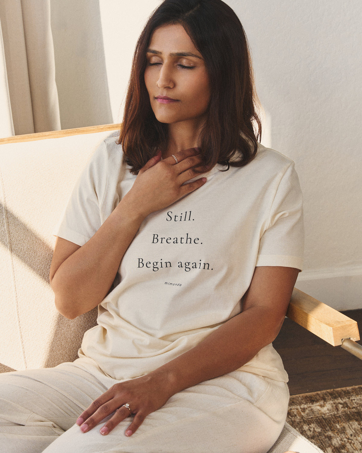 A woman with her eyes closed rests a hand on her chest, in the Still Tee. The print reads: Still. Breathe. Begin again.