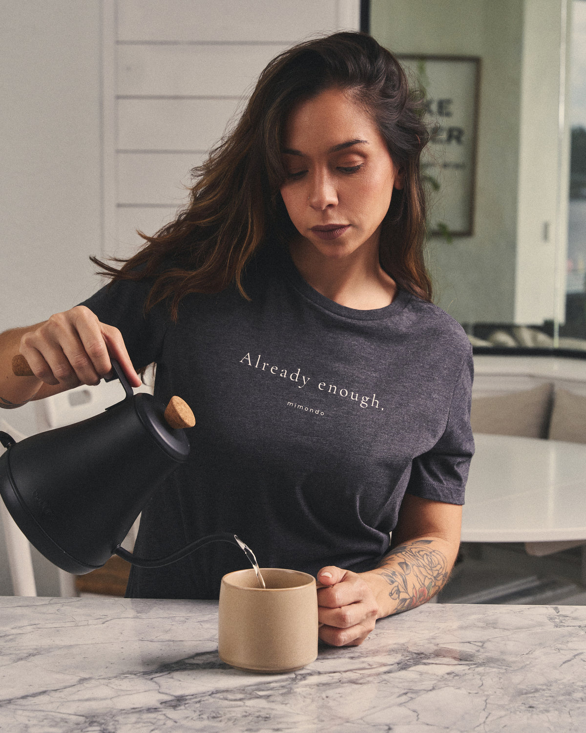 A woman pours water from a kettle into a mug at a kitchen counter, in the Strength Tee. The print reads: Already enough.