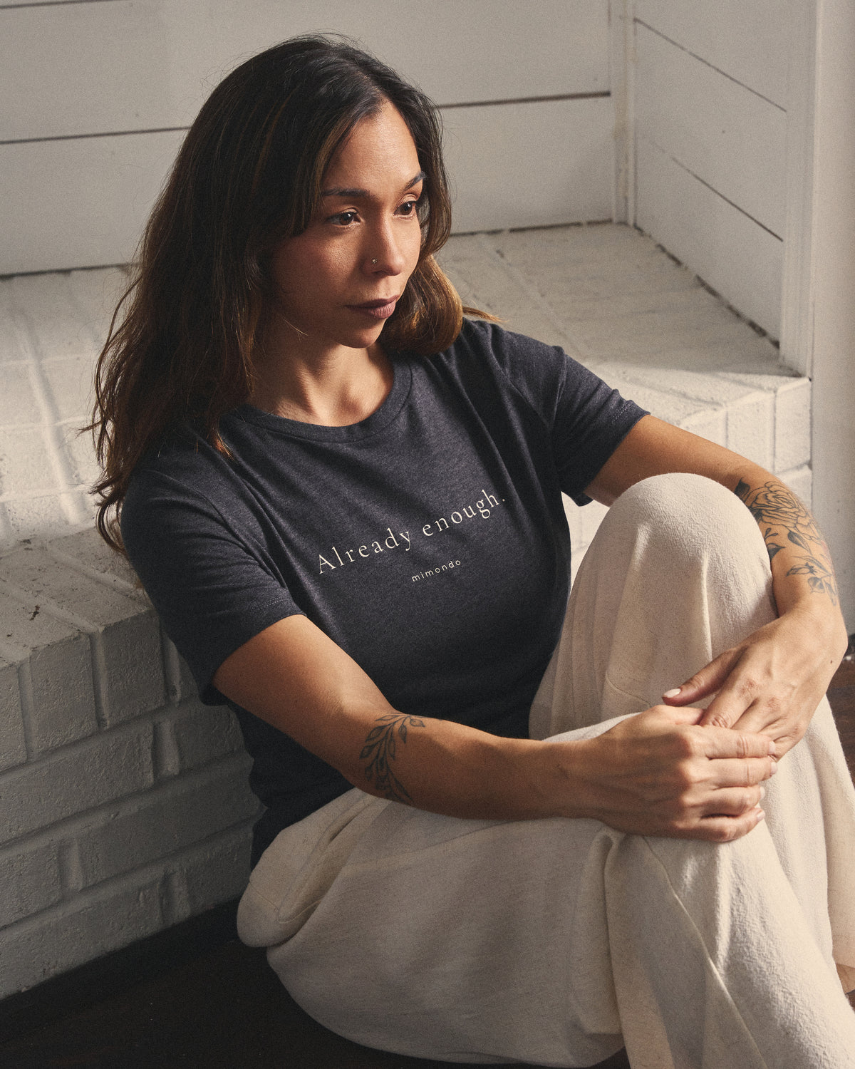 A woman sits on the floor with her arms around her knee, in the Strength Tee. The print reads: Already enough.