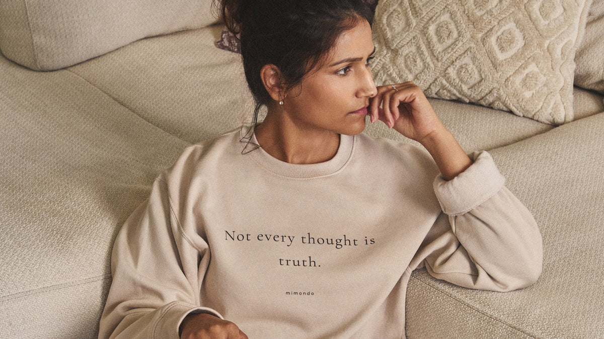 A woman resting against a sofa in the Grounded Sweatshirt. The print reads: Not every thought is truth.